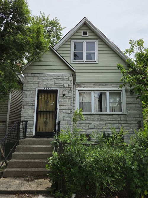Front view of a two-story house with light green siding and a stone front, concrete steps leading to a black door, and overgrown shrubs in front.