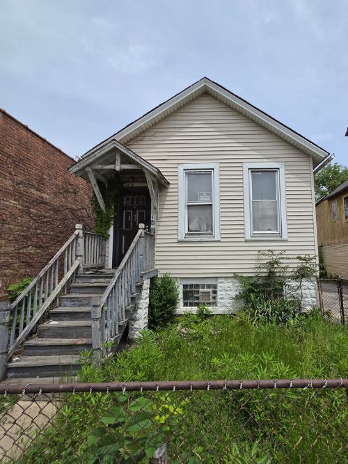 A small, weathered house with a wooden porch and overgrown yard behind a chain-link fence.