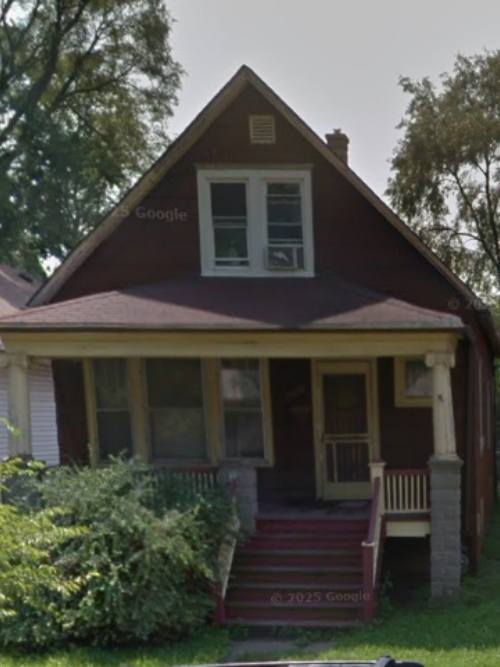 Wooden, two-story house with a front porch and red steps leading to a small entryway, surrounded by shrubs and trees.