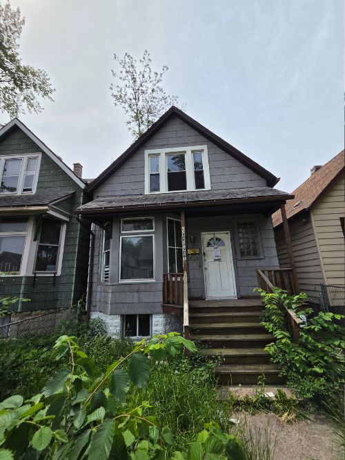 Two-story gray cottage with a small porch, white front door, and wooden steps, flanked by overgrown plants and neighboring houses.