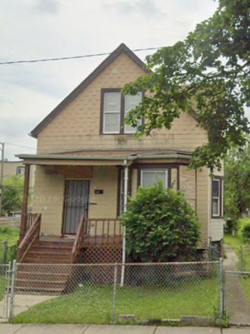 Two-story beige house with a small front porch, steps, and a chain-link fence surrounding a grassy yard.