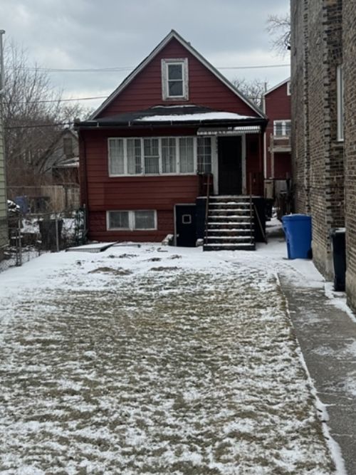 Red wooden, two-story house with a peaked roof, front stairs, and a black door; snow-dusted yard and a blue trash bin nearby.